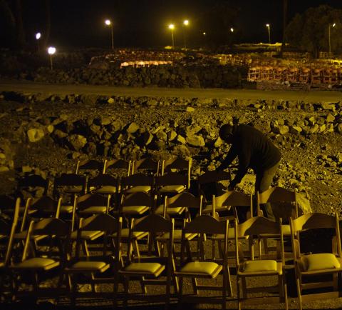 a shadowy figure leans over chairs arranged in rows in a rubble strewn nighttime outdoor setting. a shadowy figure leans over chairs arranged in rows in a rubble strewn nighttime outdoor setting.