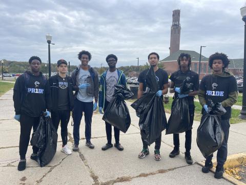 8 student athletes wearing their CSI uniform are standing in front 1 R Recreation building.​  They are holding black garbage bags filled with trash they picked up on campus.